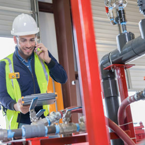 Young adult Hispanic man is an engineer working in oil and gas industry. He is inspecting pipeline equipment and making a phone call. Man is wearing hard hat and safety vest.